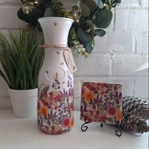 Może przedstawiać: Biały ceramiczny wazon z kwiatowym wzorem z prasowanych kwiatów w odcieniach pomarańczowego, fioletowego i zielonego. Wazon ma wąską szyjkę i szeroką podstawę. Na zdjęciu znajduje się również mała kwadratowa płytka z podobnym kwiatowym wzorem.