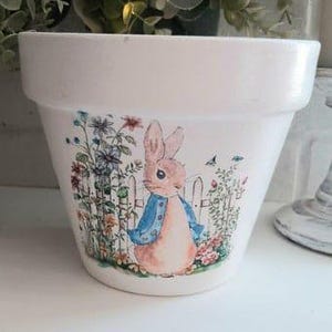 Pode incluir: Vaso de cerâmica branco com uma ilustração do Peter Rabbit. A ilustração mostra o Peter Rabbit vestindo uma jaqueta azul e em pé em um jardim com flores e uma cerca branca.