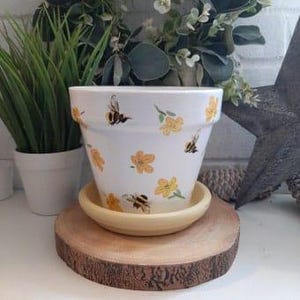Może przedstawiać: Biała doniczka ceramiczna z wzorem pszczół i kwiatów w kolorze żółtym i brązowym. Doniczka ma pasującą podstawkę.