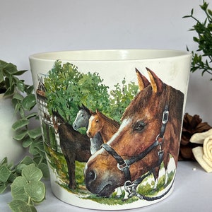 Könnte beinhalten: Weißer Keramiktopf mit einem Aquarelldesign von vier Pferden auf einer Weide. Die Pferde sind braun, grau und weiß. Der Topf hat eine abgerundete Form und einen breiten Rand.