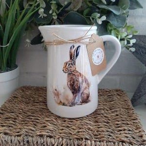 Könnte beinhalten: Weißer Keramikkrug mit Henkel, verziert mit einer detaillierten Illustration eines braunen Hasen. Der Krug hat eine Schnurschleife und ein kleines Etikett. Er steht auf einer gewebten Matte.