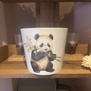 Puede incluir: Maceta de cerámica blanca con una ilustración de un panda blanco y negro sosteniendo bambú. El panda está rodeado de hojas de bambú verdes.