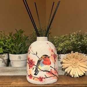 Può includere: Bottiglia diffusore in ceramica bianca con un design di pettirosso e bacche rosse. I bastoncini diffusori neri sono nella bottiglia.