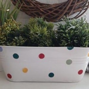 Op de afbeelding: Witte metalen plantenbak met een stippenpatroon in rood, groen, geel en blauw. De plantenbak is ovaalvormig en heeft een licht verweerde afwerking.