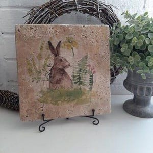 Puede incluir: Una baldosa cuadrada de cerámica con un conejo marrón rodeado de flores y helechos verdes y amarillos. La baldosa está sobre un soporte de metal negro.