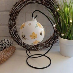 May include: White ceramic Christmas ornament with a bee and yellow flower design. The ornament is hanging from a black metal stand.