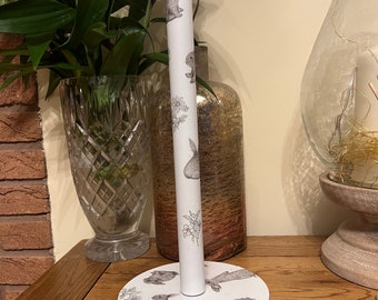 May include: White metal paper towel holder with a silver top. The holder is decorated with a pattern of bunnies and flowers.