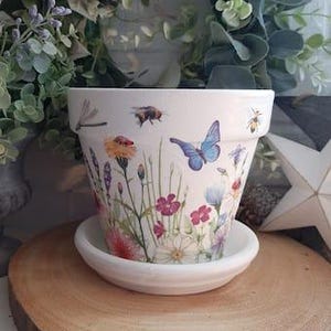 Op de afbeelding: Een witte keramische bloempot met bijpassende schotel. De pot is versierd met een kleurrijk bloemen- en insectenontwerp, waaronder vlinders, bijen en wilde bloemen. De pot staat op een rond houten oppervlak.