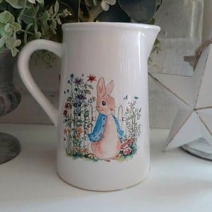 Può includere: Brocca in ceramica bianca con un'illustrazione di Peter Rabbit. La brocca presenta un coniglio vestito di blu in piedi in un giardino con fiori e una staccionata bianca.