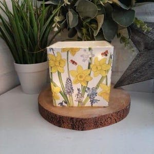 Op de afbeelding: Een vierkante witte keramische kaarsenhouder met een bloemenontwerp met gele narcissen, blauwe hyacinten en witte bloemen. De kaarsenhouder heeft een lichtje erin, waardoor het bloemenontwerp wordt verlicht.