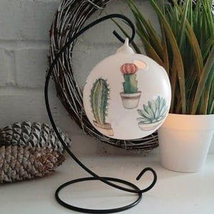 Puede incluir: Adorno de cerámica blanca con un soporte de metal negro. El adorno presenta un diseño pintado de cactus y suculentas en macetas.
