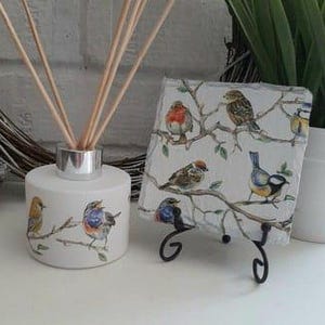 May include: A white ceramic essential oil diffuser with a silver top and wooden reeds, decorated with birds. A matching bird-themed coaster is on a black metal stand. The items are on a white surface.