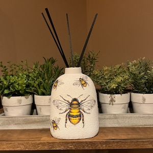May include: White ceramic diffuser bottle with a bee design. The bottle has black diffuser sticks and is decorated with multiple bees in yellow and black.