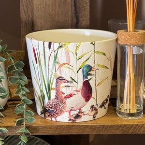 May include: White ceramic planter with a duck family design. The planter features a cracked finish and a detailed illustration of ducks, reeds, and cattails. The planter is cylindrical and sits on a wooden shelf, suitable for indoor or outdoor use.