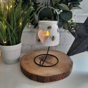 May include: White ceramic candle holder with a heart-shaped cutout and a bee design. The holder has a metal ring for hanging and is on a black metal stand.