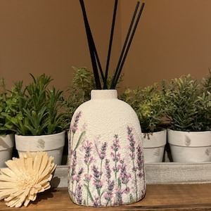 May include: White ceramic diffuser bottle with a lavender floral design and black diffuser sticks. The bottle is sitting on a wooden shelf.