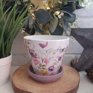 Peut inclure: Un pot de fleurs en céramique blanche avec un motif floral dans des tons de rose, violet, jaune et vert. Le pot a une soucoupe assortie de couleur violet clair.