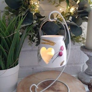 May include: White ceramic candle holder with a heart-shaped cutout and floral design. The holder is on a white metal stand with a round base.