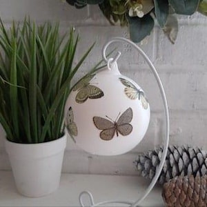 Könnte beinhalten: Weißer Glaskugelschmuck mit einer Aufhängeschlaufe und einem weißen Metallständer. Der Kugelschmuck ist mit einem Muster aus braunen, grünen und gelben Schmetterlingen verziert.