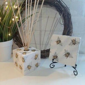 Op de afbeelding: Een witte houten kubusdiffuser met een bijenpatroon en een witte keramische tegel met een bijenpatroon op een zwarte standaard.