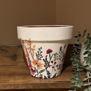 Puede incluir: Maceta de flores de terracota blanca con un patrón floral en tonos naranja, rosa, morado y verde.