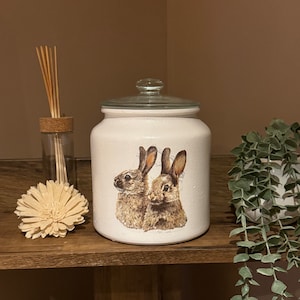 以下が含まれることがあります： 白いセラミック製のジャーにガラスの蓋が付いており、2羽の茶色のウサギが描かれたデザインが施されています。ジャーは、小さな白い花とスティックが入ったガラスの花瓶が置かれた木製の棚の上にあります。