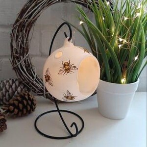 May include: White ceramic candle holder with a bee design. The holder is hanging from a black metal stand.