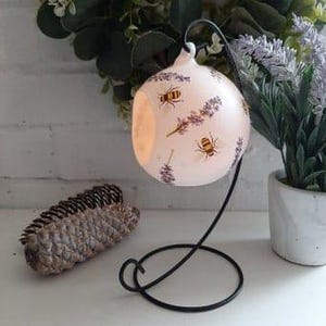 以下が含まれることがあります： 蜂とラベンダーの花柄が描かれた、カットアウトデザインの白いセラミック製キャンドルホルダー。ホルダーは黒い金属製のスタンドの上に置かれています。