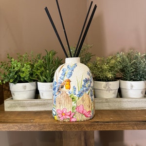 Puede incluir: Botella difusora de cerámica blanca con un diseño floral y un pájaro. La botella tiene una superficie texturizada y está llena de varillas difusoras negras.