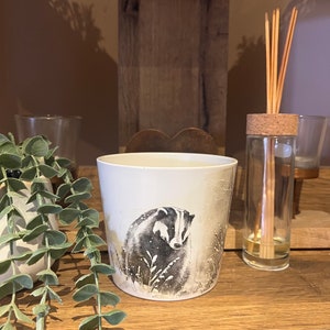 Puede incluir: Portavelas de cerámica blanca con una ilustración en blanco y negro de un tejón en un paisaje de bosque nevado.