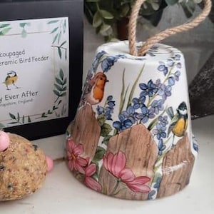 Könnte beinhalten: Ein Keramik-Vogelhäuschen in Glockenform mit einem Seilgriff. Der Futterspender ist mit einem weißen Hintergrund und bunten Blumen, Vögeln und Holzlatten verziert. Der Text "Happy Ever After" ist auf der Schachtel aufgedruckt.