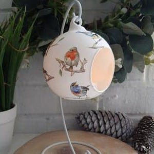 May include: White ceramic candle holder with a cut-out design. The holder is decorated with a pattern of colorful birds on branches. It is hanging from a metal stand.