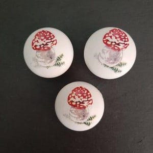 May include: Three white ceramic knobs with a red and white mushroom design. Each knob features a red mushroom with white spots on a white background with green leaves.