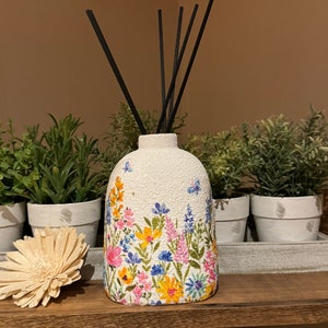 Op de afbeelding: Witte keramische diffuserfles met een bloemenontwerp met roze, gele en blauwe bloemen. Zwarte diffuserstokjes zijn in de fles gestoken.