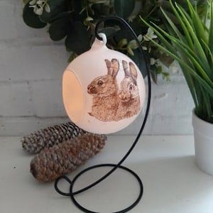 May include: White ceramic hanging ornament featuring a brown rabbit design. The ornament has a hole in the side for a tea light candle. The ornament is suspended from a black metal stand.