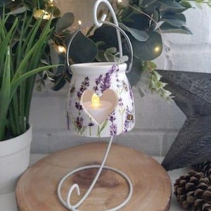 May include: White ceramic candle holder with a lavender and bee design. The holder has a heart-shaped cutout and is on a metal stand.