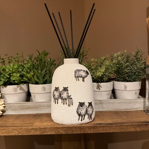 May include: White ceramic diffuser with a textured finish, decorated with black and white sheep designs. The diffuser contains black reeds. It is displayed on a wooden shelf. Small potted plants are in the background.