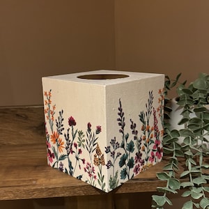 Könnte beinhalten: Ein weißer, hölzerner Taschentuchbox-Bezug mit einem Blumendesign. Das Design zeigt eine Vielzahl von bunten Blumen, darunter rosa, lila, orange und gelb.