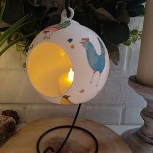 May include: White ceramic candle holder with a cut-out design and a black metal stand. The holder features a colorful pattern of birds and flowers.