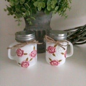 May include: Two white ceramic jars with pink floral designs and silver lids. The jars have handles and are tied with twine.