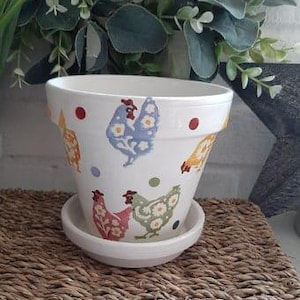 Puede incluir: Maceta de cerámica blanca con un patrón de pollo en tonos de azul, amarillo, rojo y verde. La maceta tiene un platillo a juego y presenta un patrón de lunares.