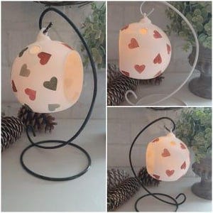 May include: White ceramic candle holder with a hanging loop and a black metal stand. The holder is decorated with red and green hearts.