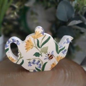 Könnte beinhalten: Eine weiße Keramik-Teekanne mit einem Blumenmuster mit gelben und blauen Blumen, grünen Blättern und einer Biene. Die Teekanne ist ein Dekorationsobjekt und nicht funktionsfähig.