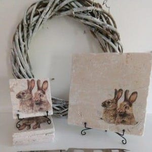 Puede incluir: Conjunto de cuatro posavasos cuadrados de cerámica con una imagen de dos conejos en marrón y blanco. Los posavasos se muestran en un soporte blanco.