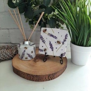 May include: A white ceramic diffuser bottle with a lavender and bee design and a matching square diffuser reed stand. The diffuser bottle has a silver cap and is filled with reeds.