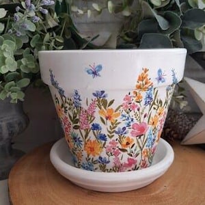 May include: White ceramic flower pot with a colorful floral design and butterflies. The pot has a matching saucer.