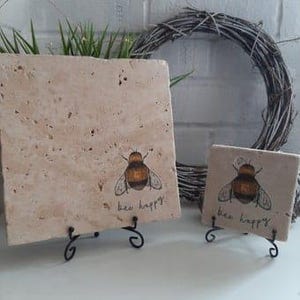 Puede incluir: Dos posavasos cuadrados de cerámica beige con un soporte de metal negro. Cada posavasos presenta una ilustración de abeja negra y amarilla y el texto "bee happy".