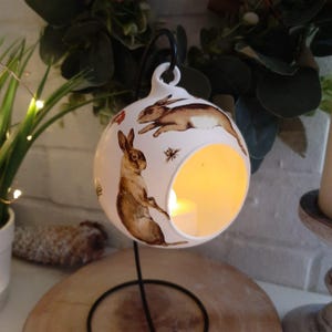 May include: A white ceramic orb candle holder with a black metal stand. The holder is decorated with brown rabbit and insect illustrations. A warm light glows from inside, creating a cosy atmosphere. The candle holder is a decorative home accent.
