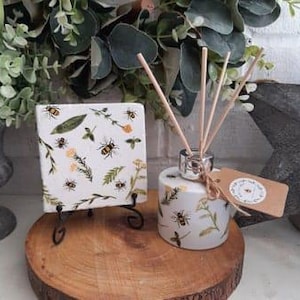 Op de afbeelding: Een witte keramische diffuserfles met een zilveren deksel en houten stokjes. De fles heeft een bloemmotief met bijen en bladeren. De diffuser staat op een kleine zwarte standaard. Er is een bijpassende keramische tegel met hetzelfde ontwerp op een aparte standaard.