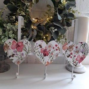 May include: Three white metal heart-shaped hooks with a floral and bee design. The hooks are attached to a white metal bar.